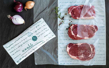 Einschlagpapier das auf einem Tisch ausgebreitet ist. In dem Einschlagpapier liegen Steaks die verapckt werden sollen